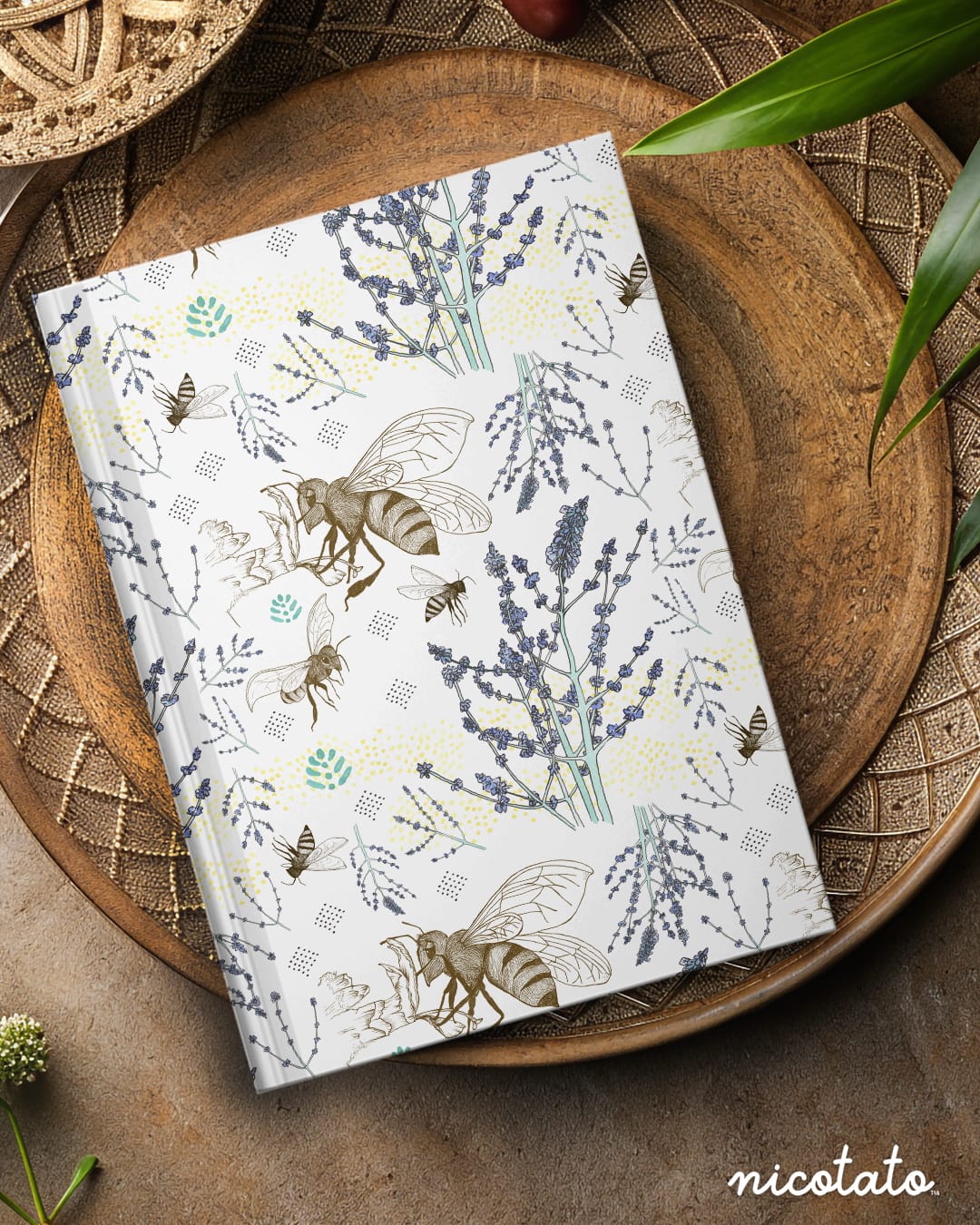 A hardcover notebook with a botanical and bee pattern, displayed on a round wooden tray with decorative leaves and woven elements.